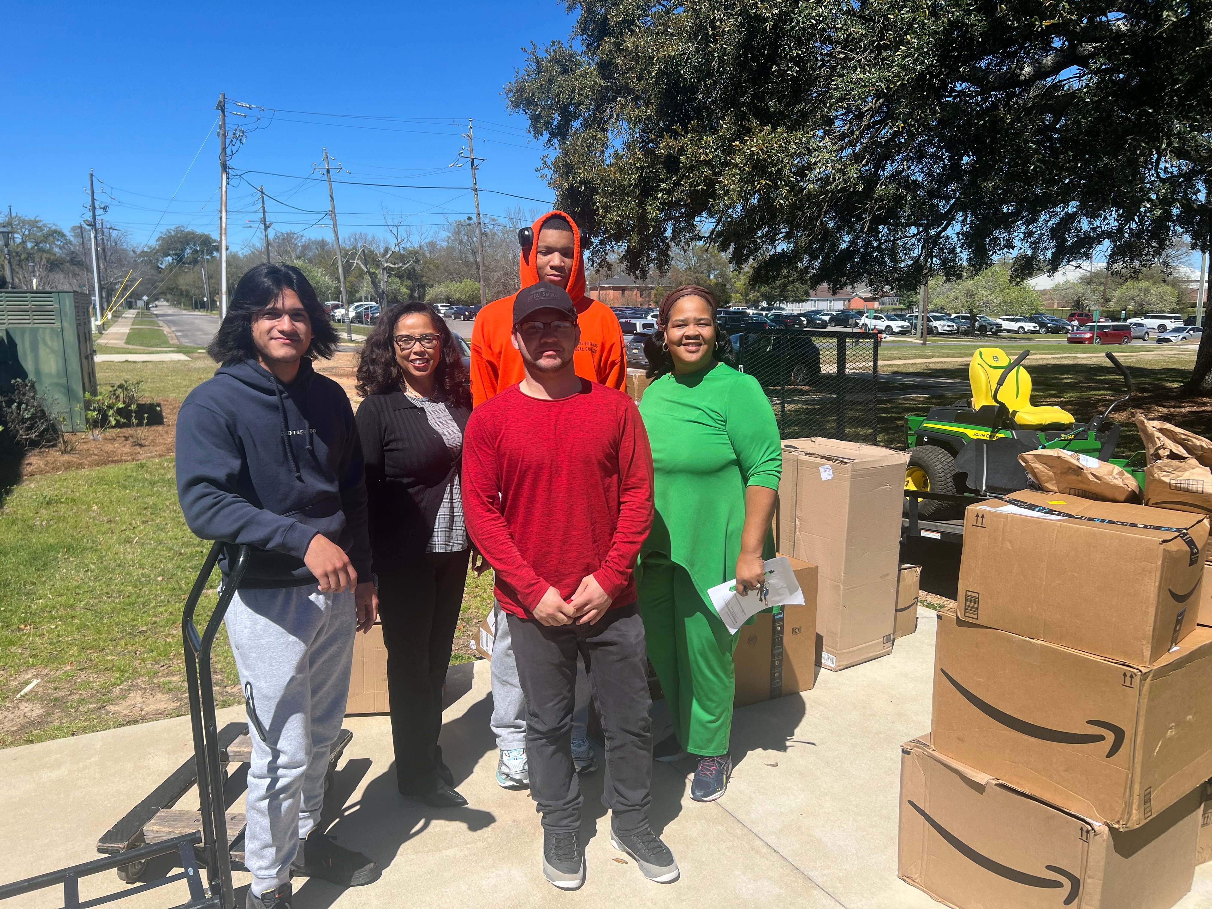 Volunteers receiving donated garden supplies alongside shipping boxes and a John Deere mower